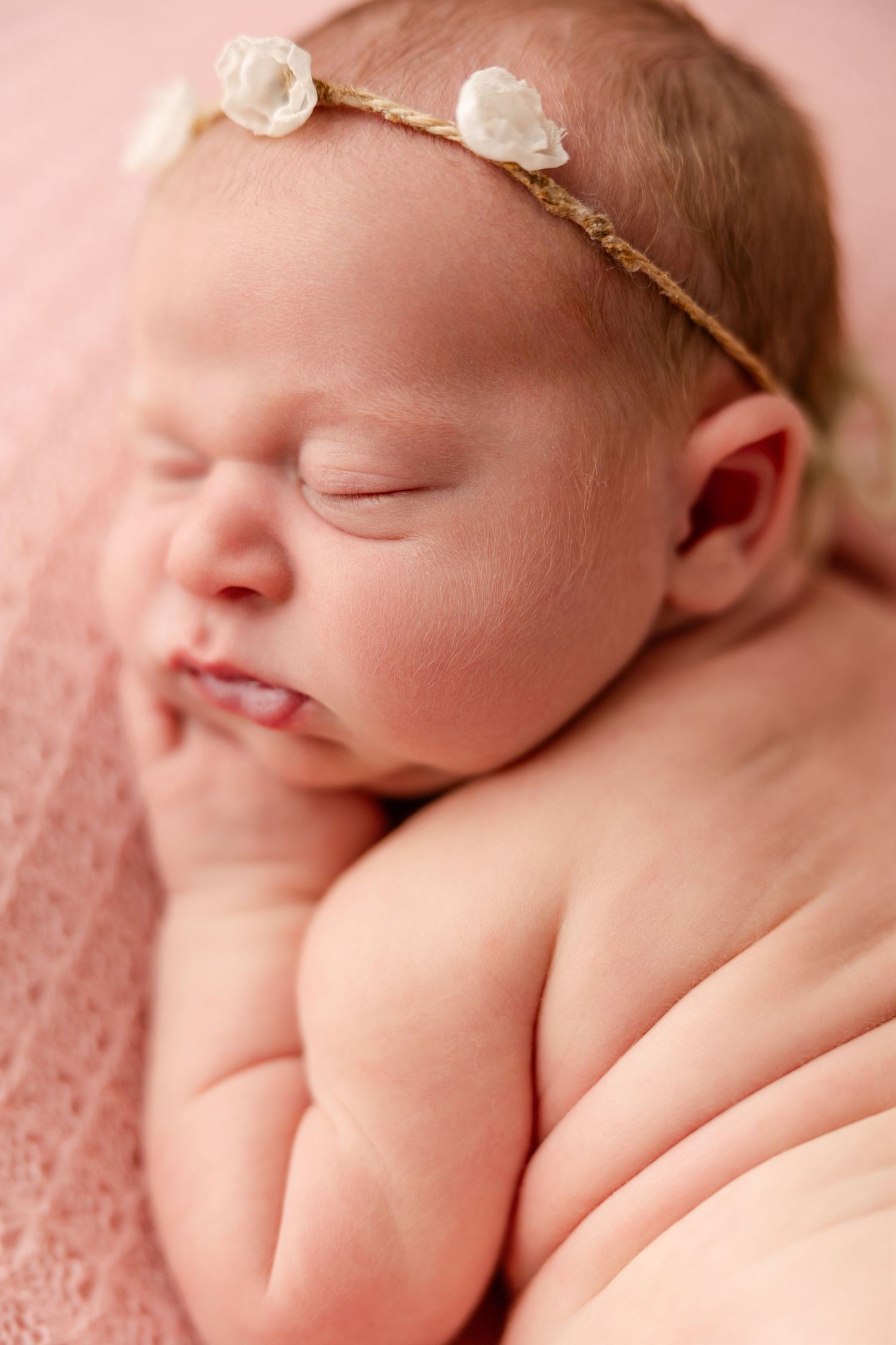 A sleeping newborn baby wearing a delicate gold headband rests peacefully against a soft pink background.