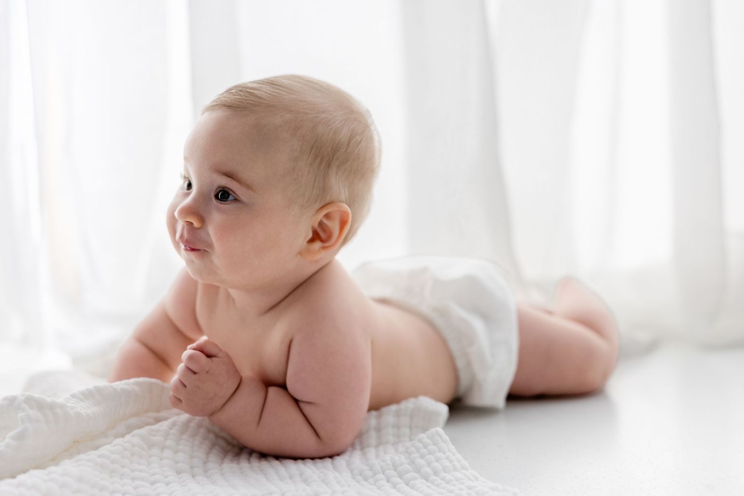 A baby lies on white bedding in a bright room with natural light streaming through sheer curtains