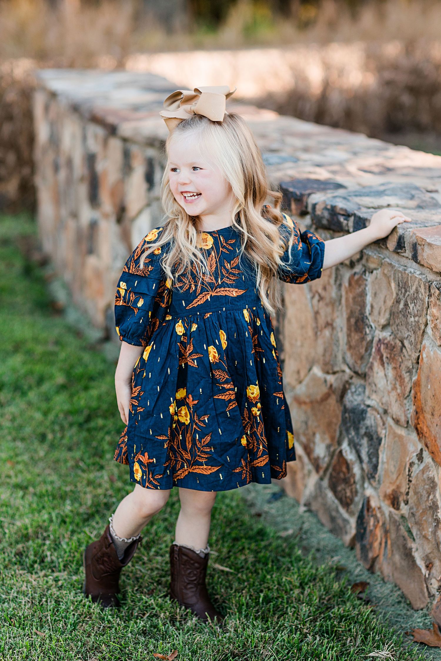 Young child in navy patterned dress sitting by stone wall outdoors.