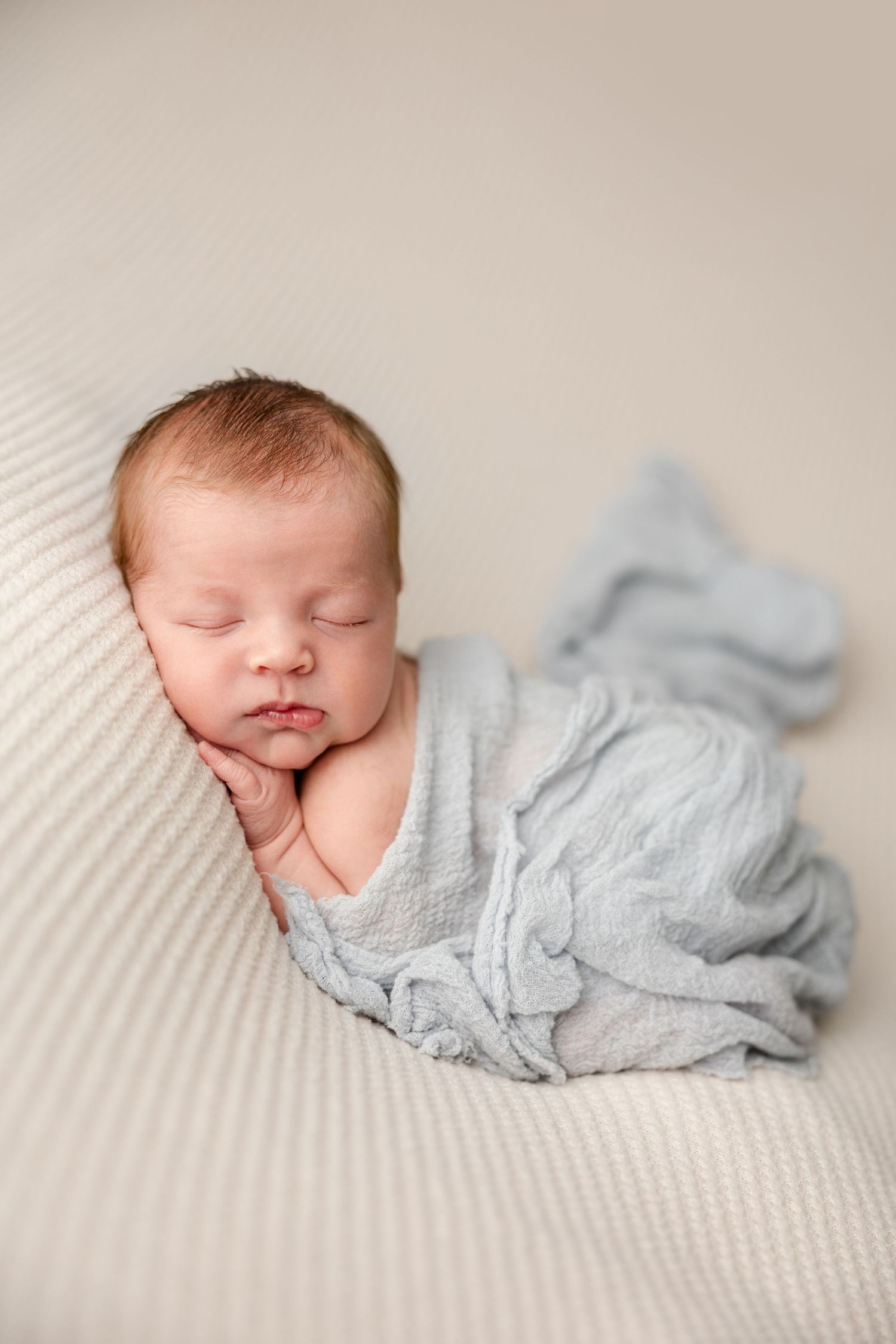 Sleeping newborn baby wrapped in soft gray fabric lying on textured white blanket.