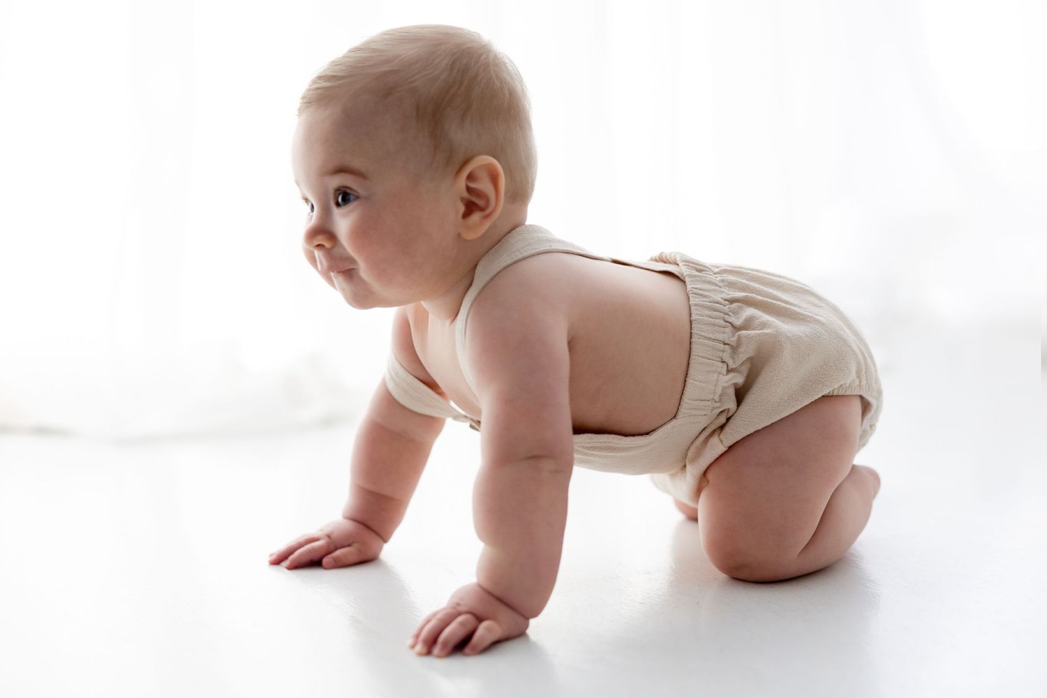 Baby in beige romper crawling on white floor during a studio photoshoot.