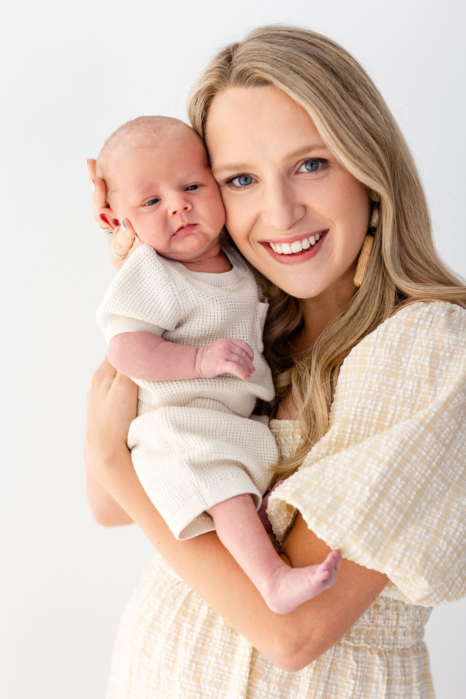 A parent in a cream sweater holds and snuggles with a baby wearing a white onesie against a white background.