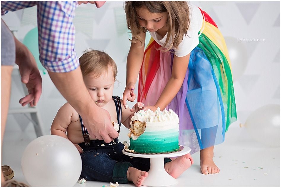 Baby celebrates first birthday with cake smash surrounded by balloons and rainbow decor.