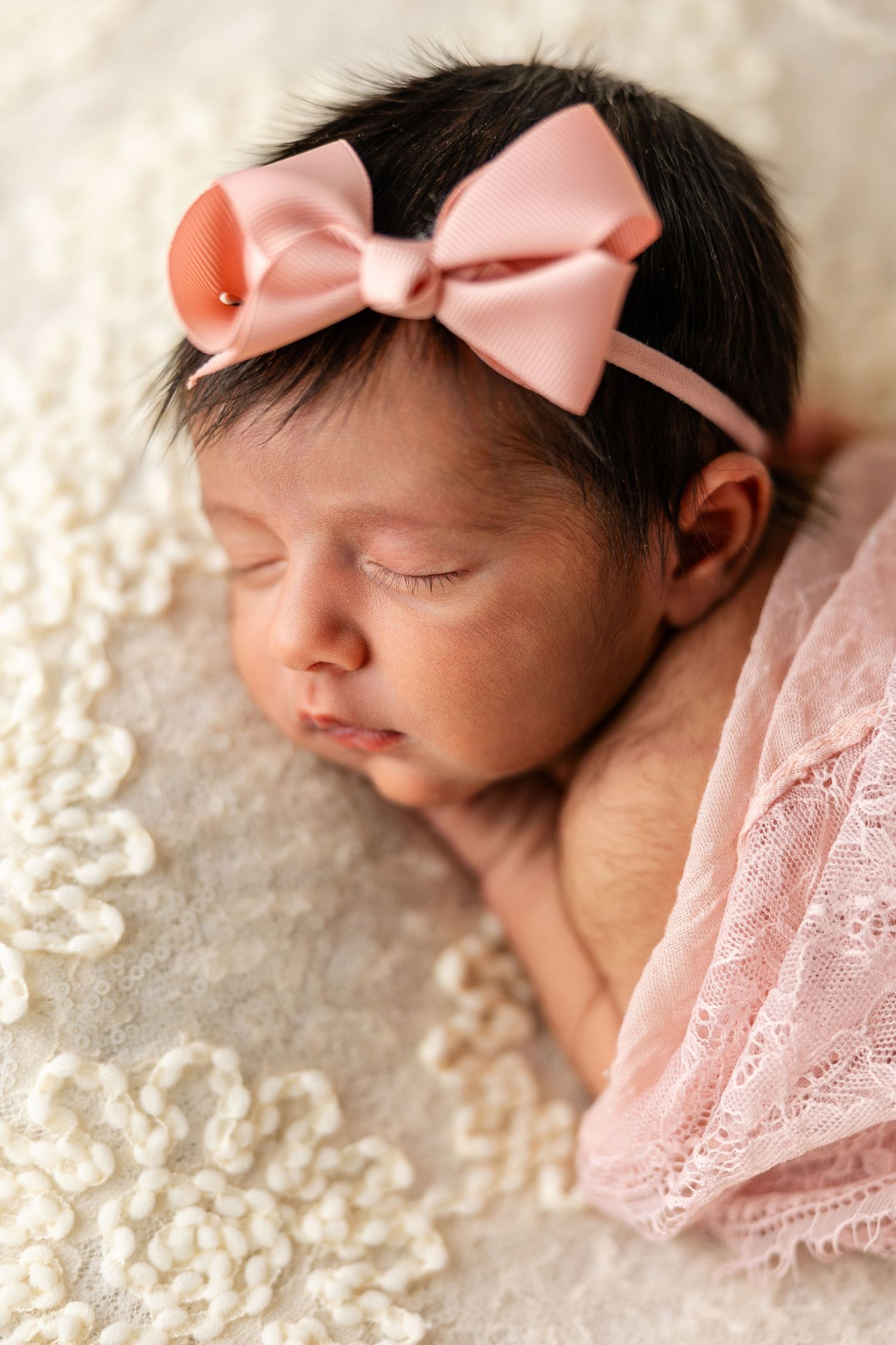 Sleeping newborn wearing pink bow headband photographed on lace fabric in soft lighting.