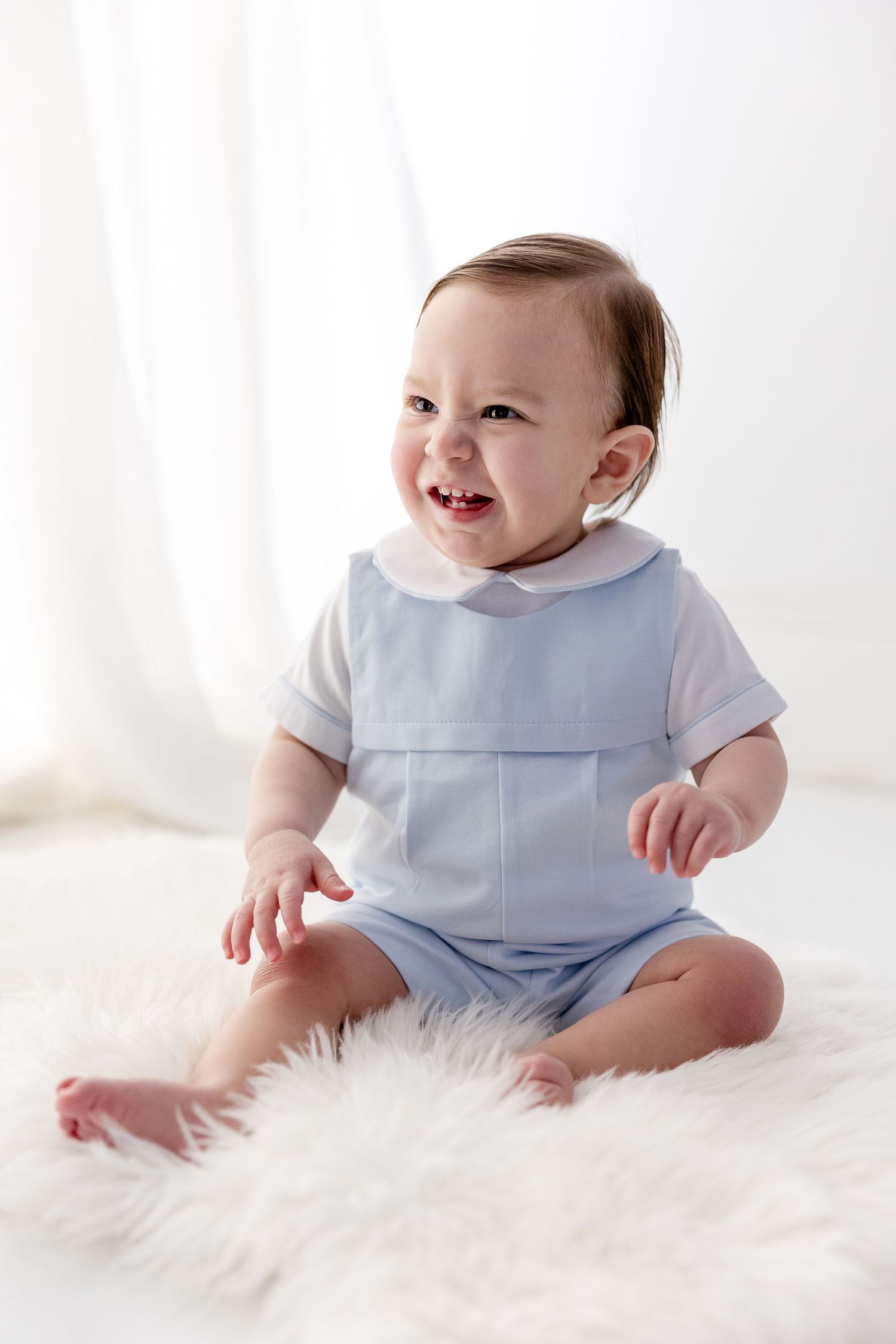 Smiling baby in pale blue outfit sits on white fluffy rug during professional photo session.