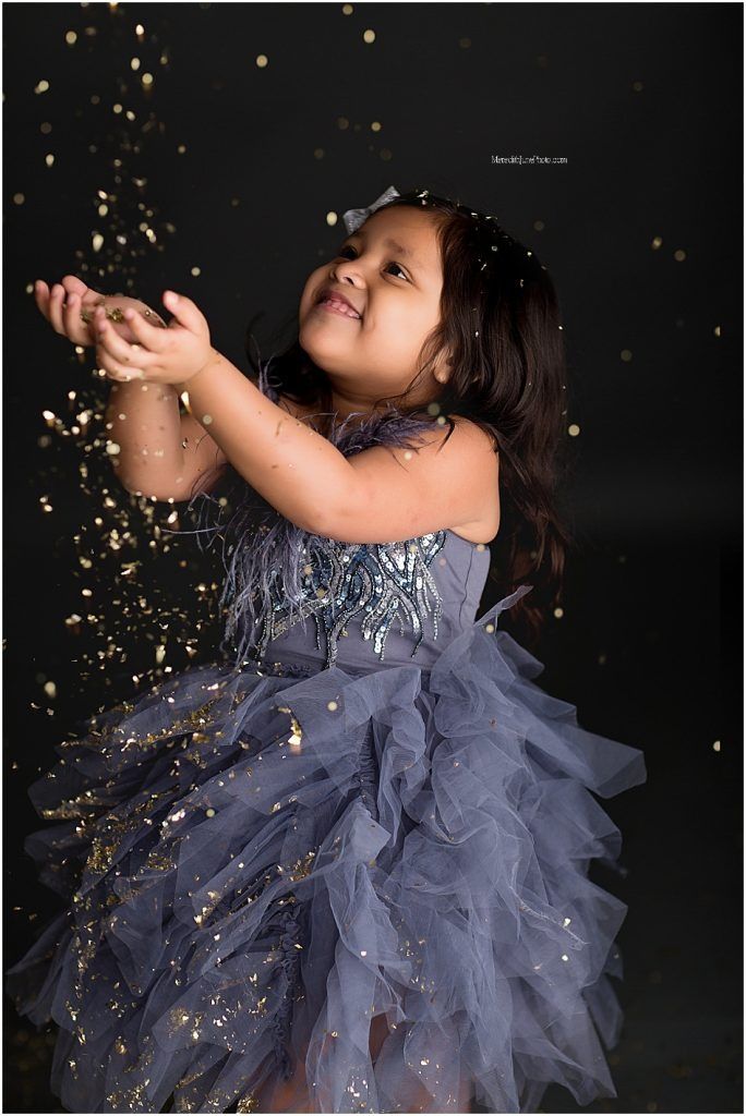 Young child in sparkly gray tulle dress catching glitter falling from above against dark backdrop.