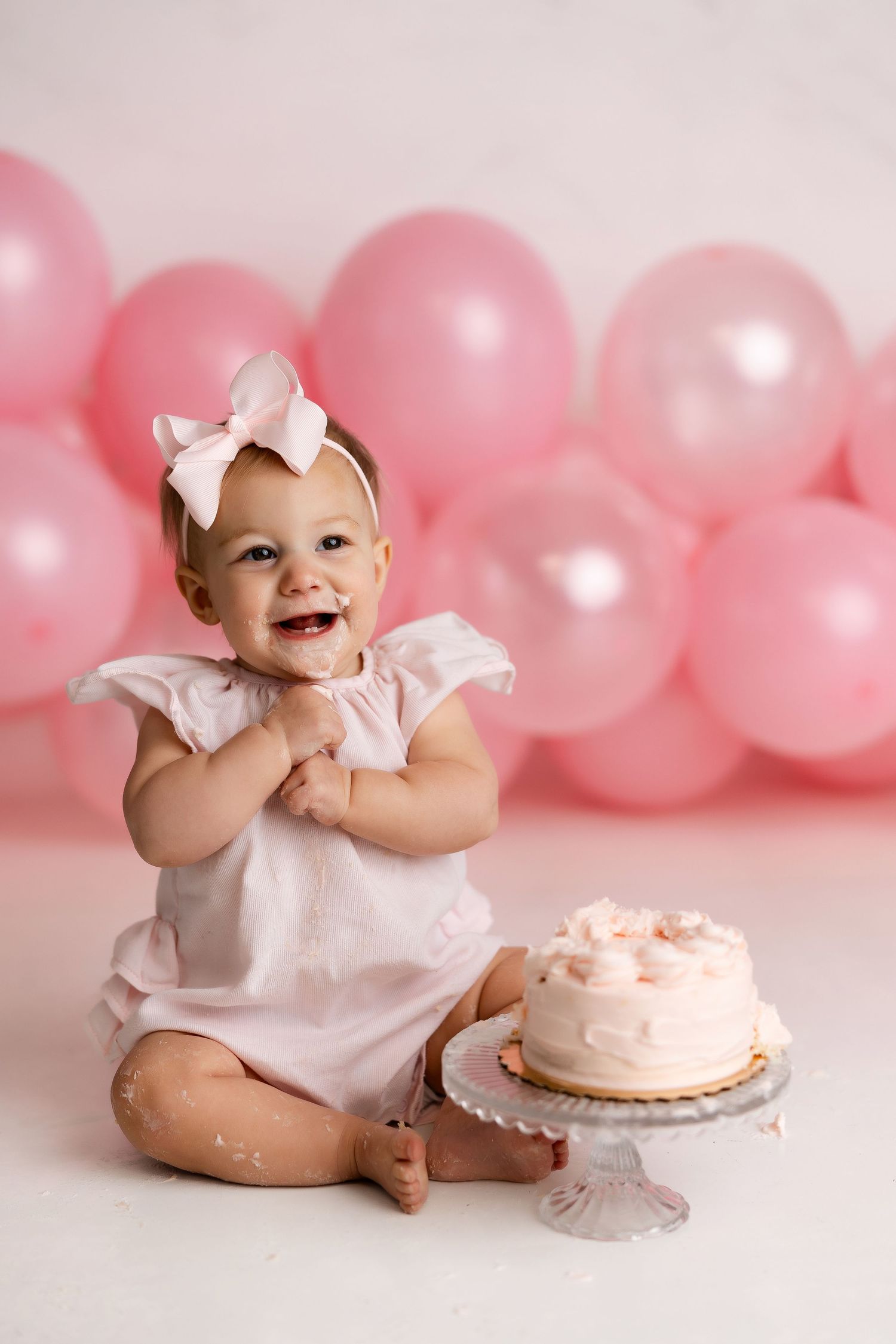 A baby in white dress sits beside pink balloons and a birthday cake during a cake smash photo session.