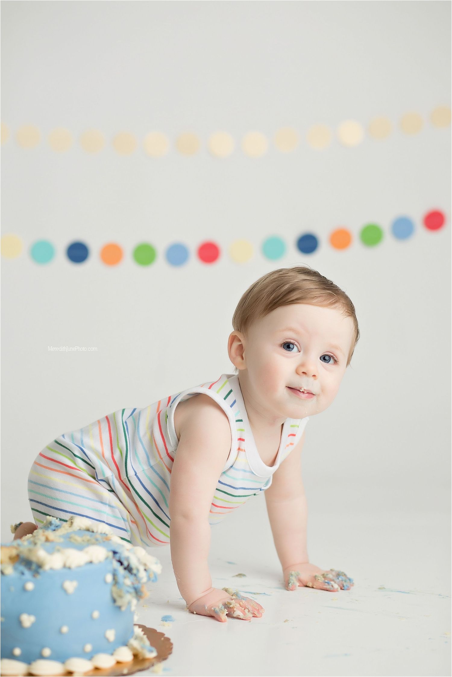 A baby in a striped outfit crawls on a white floor with colorful party decorations and a blue cake in the background.