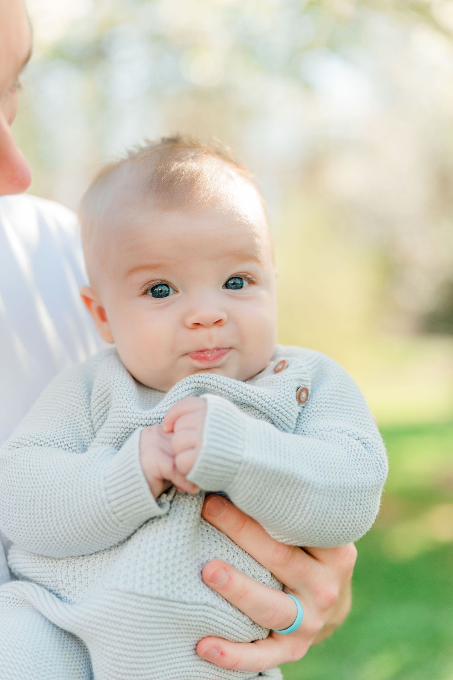 Baby in soft mint sweater makes adorable expressions while being held outdoors on sunny spring day.