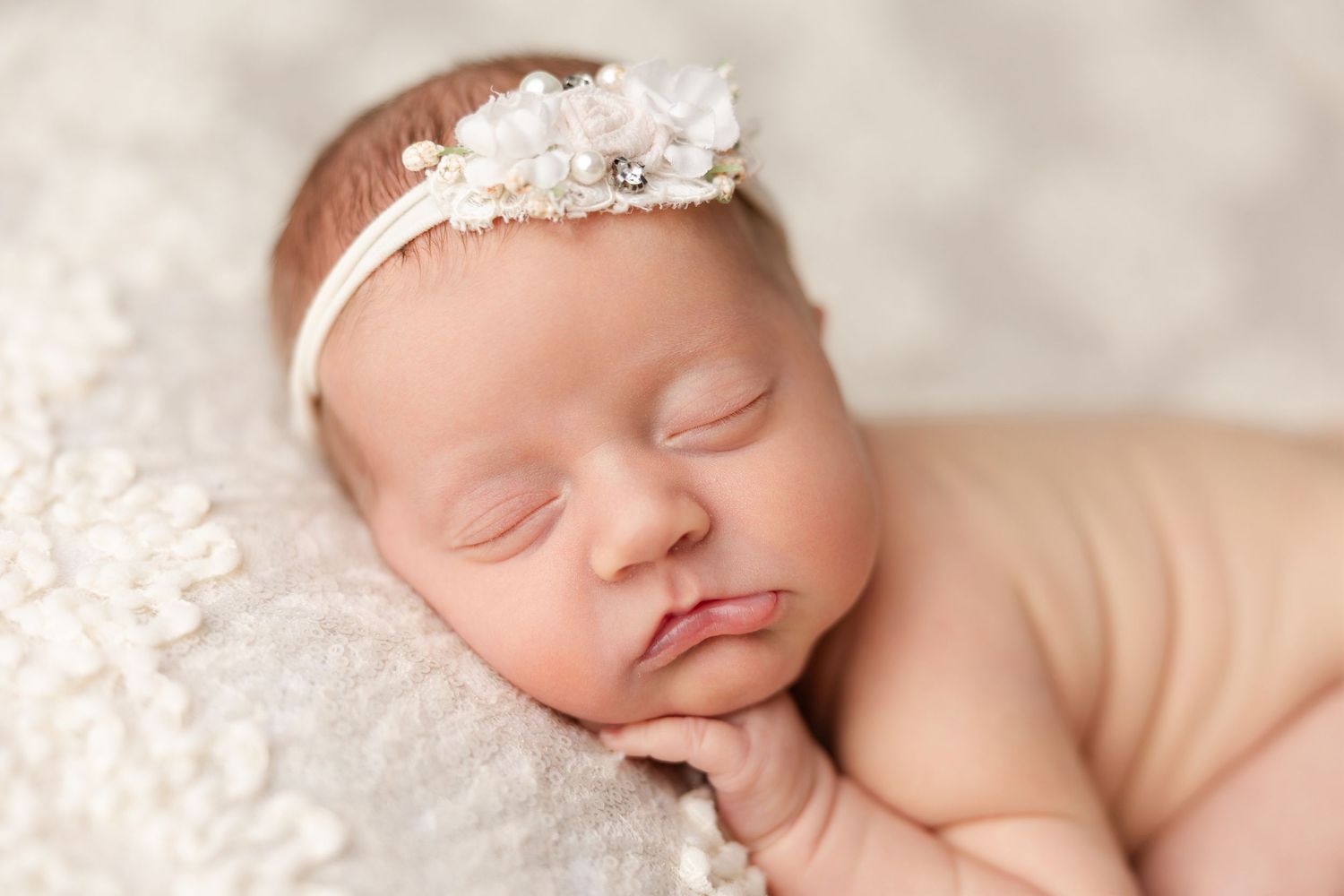 Peaceful sleeping newborn baby wearing delicate white lace headband rests on soft textured blanket.