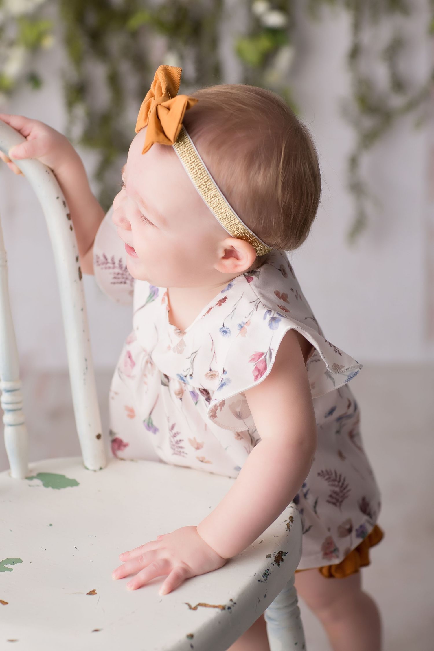 Baby in floral outfit sits by rustic white furniture with greenery hanging from above.