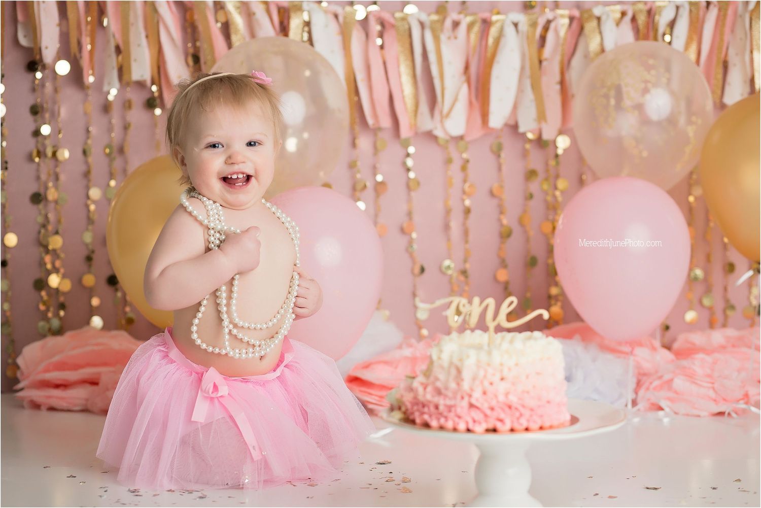 Pink and gold first birthday party decorations with tulle, balloons, pearls and cake in a festive setup.