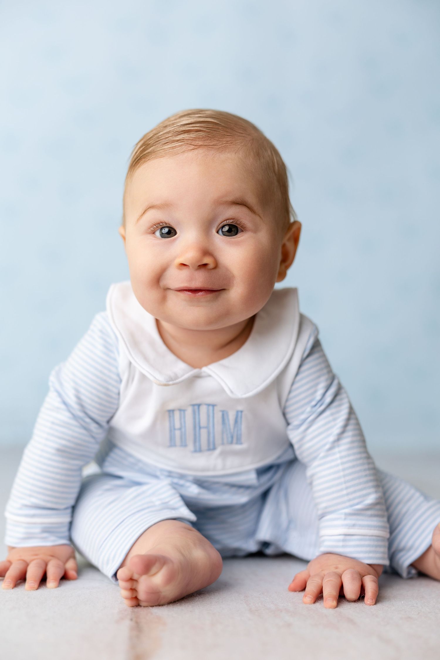 A baby in a light blue and white striped romper with a peter pan collar sits smiling against a blue background.