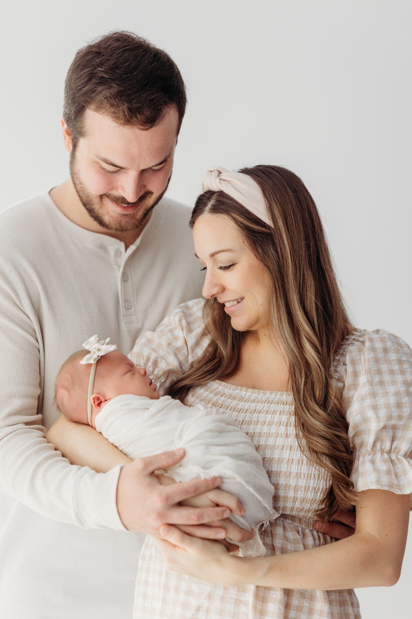 Parents in neutral colors holding newborn baby in white against light background.