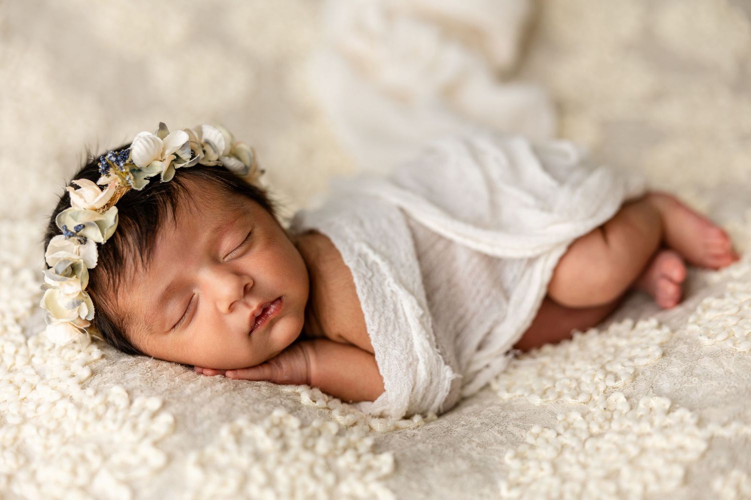 A sleeping newborn baby wearing white swaddle wrap rests peacefully on a textured cream-colored blanket.