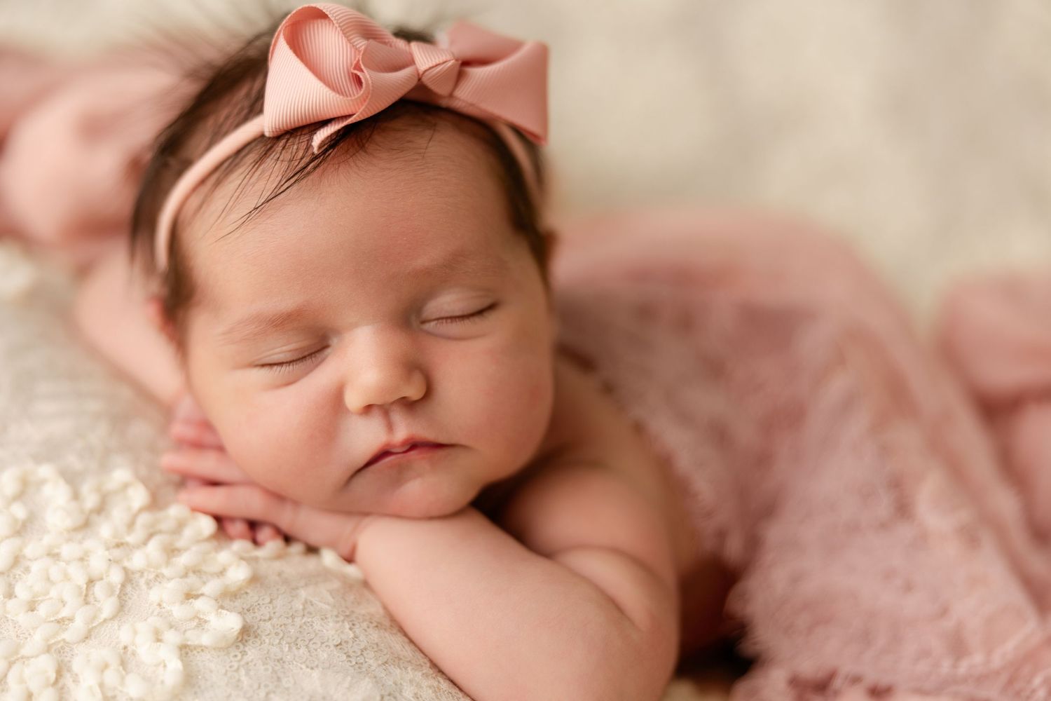 Sleeping newborn baby girl wearing a pink bow headband rests her cheek on her hands atop a cream blanket with soft pink lace.