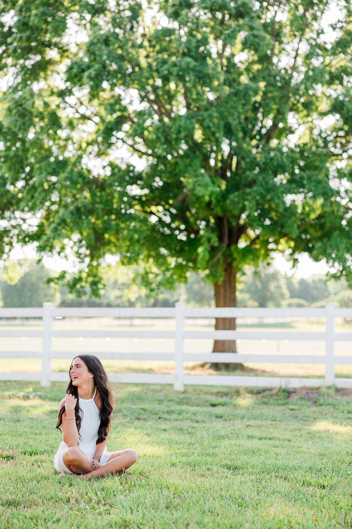 Brunette woman in white dress sitting cross-legged on grass near white fence and large tree in summer.