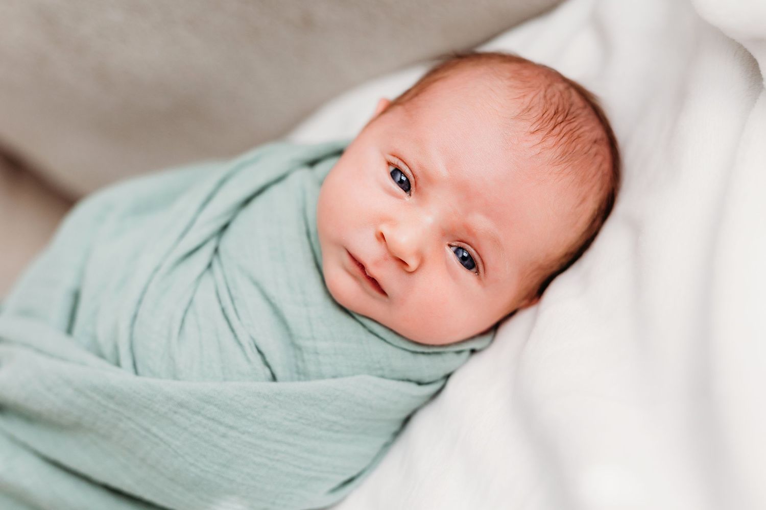 A small baby wrapped in a mint green muslin swaddle blanket rests on white bedding.