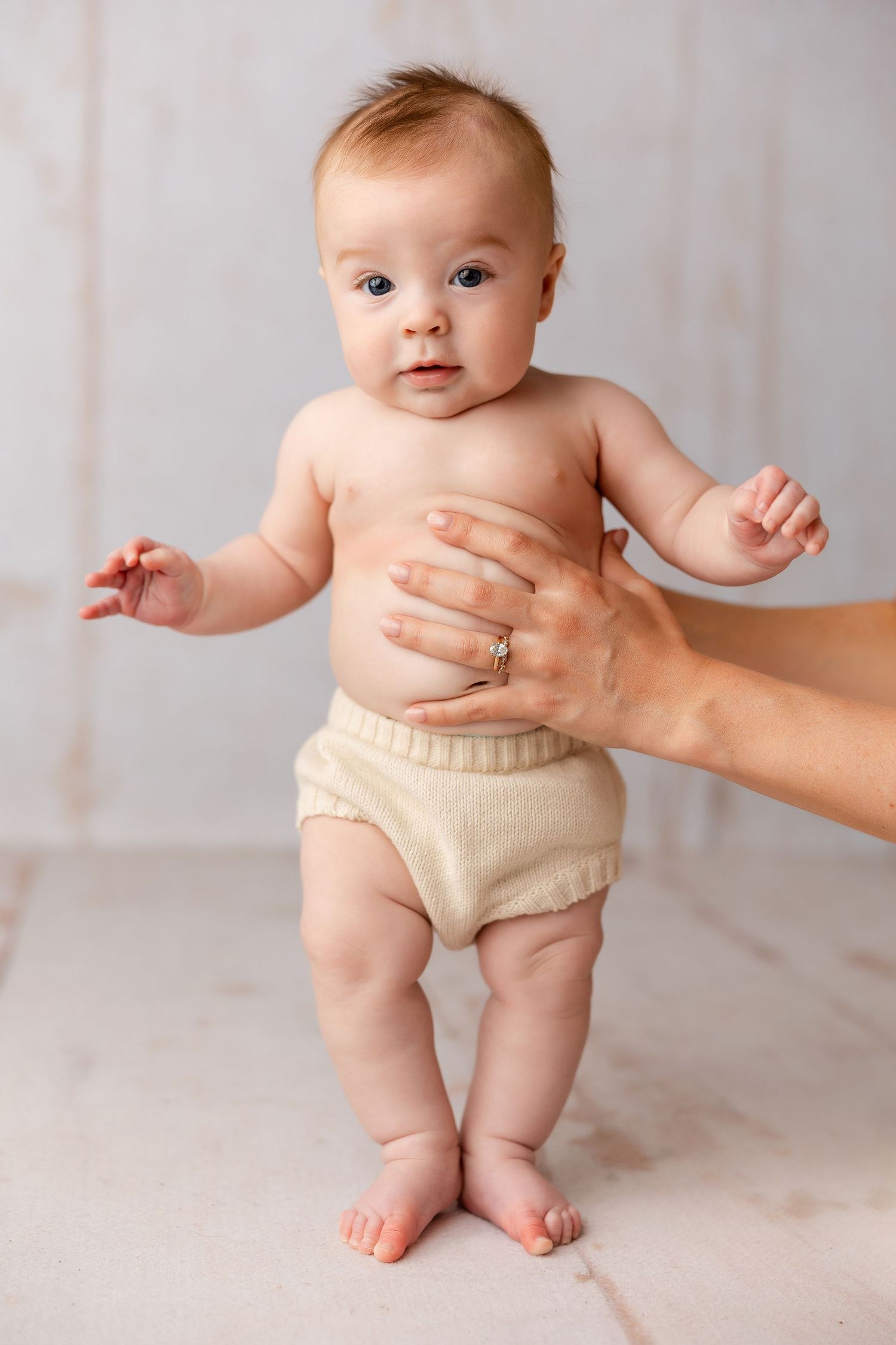 Baby learning to stand and balance in a diaper against white studio backdrop.