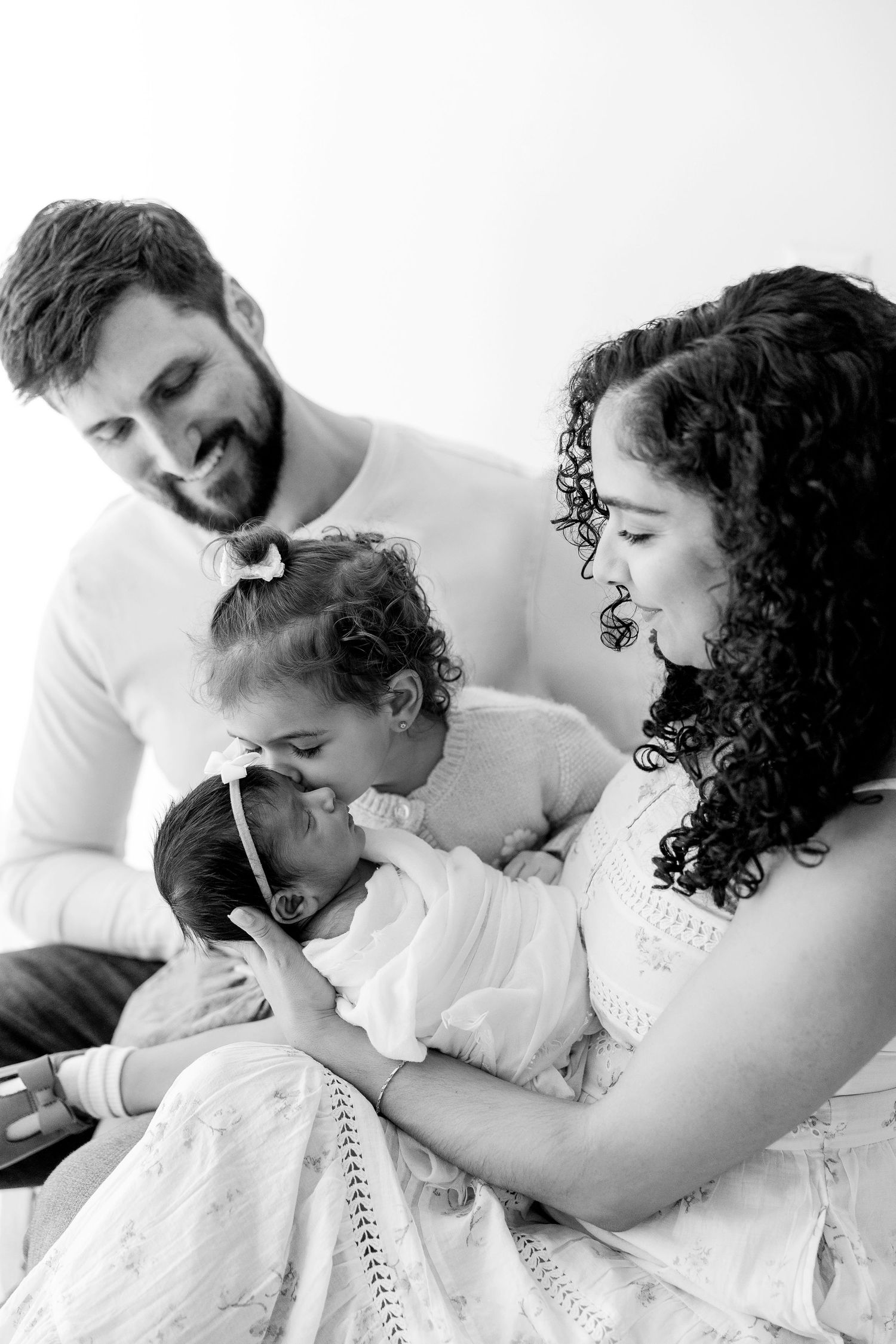 Black and white intimate family portrait showing tender moment between parents and baby in white clothing.