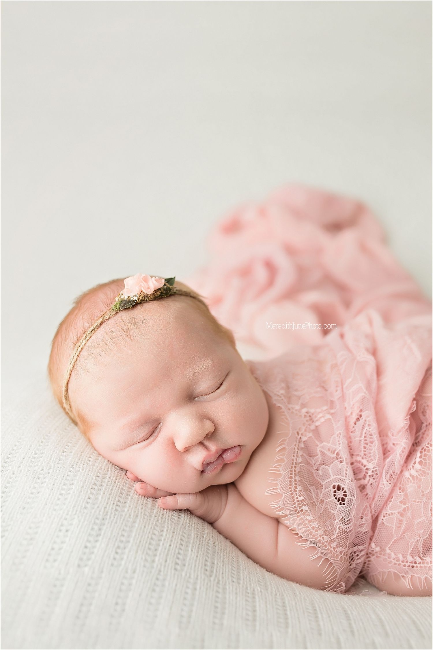 Peaceful sleeping newborn wrapped in delicate pink lace fabric on white textured surface with dainty floral headband.