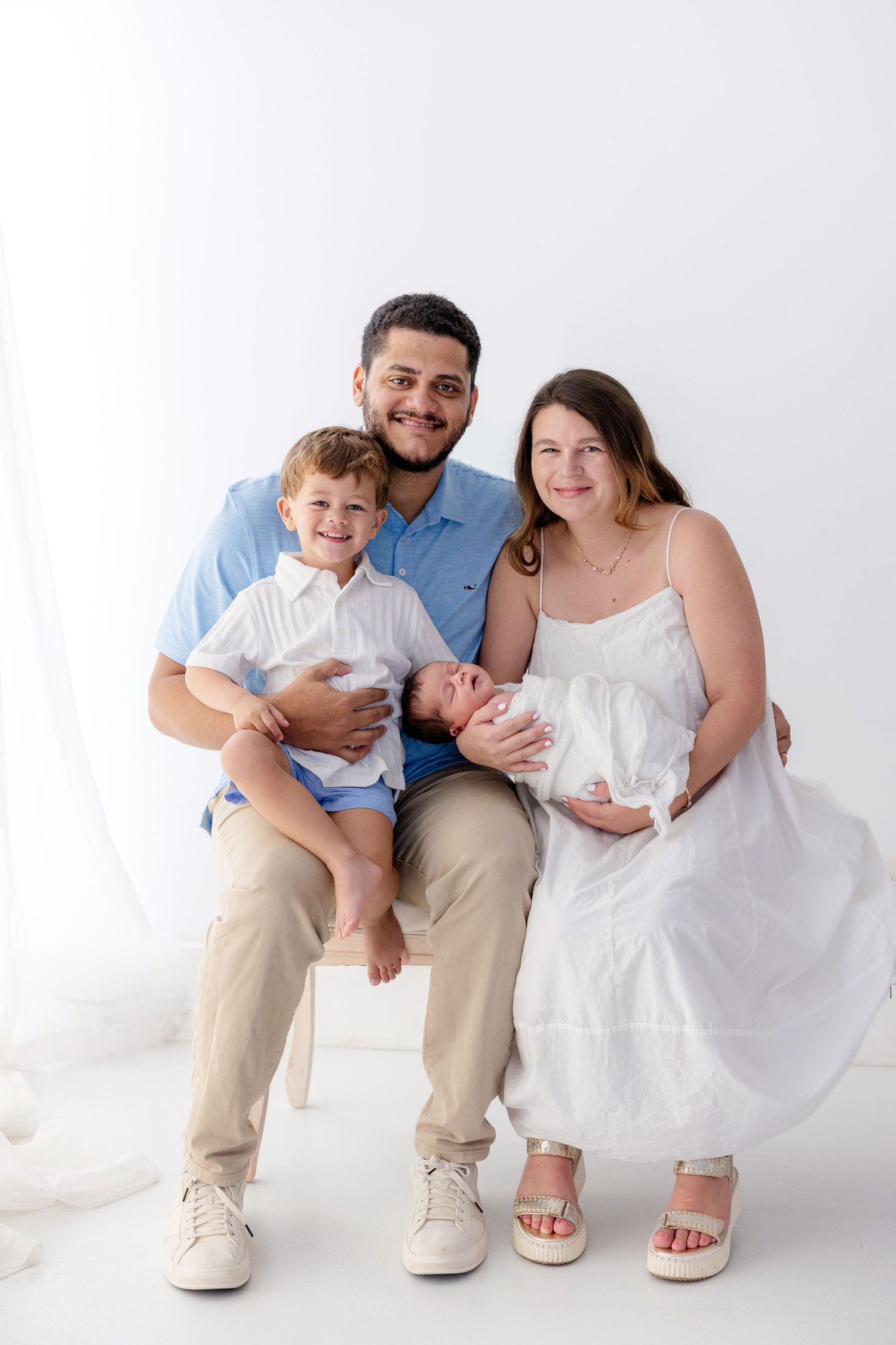 Happy family of four posing in white studio, parents holding newborn and toddler son smiling.