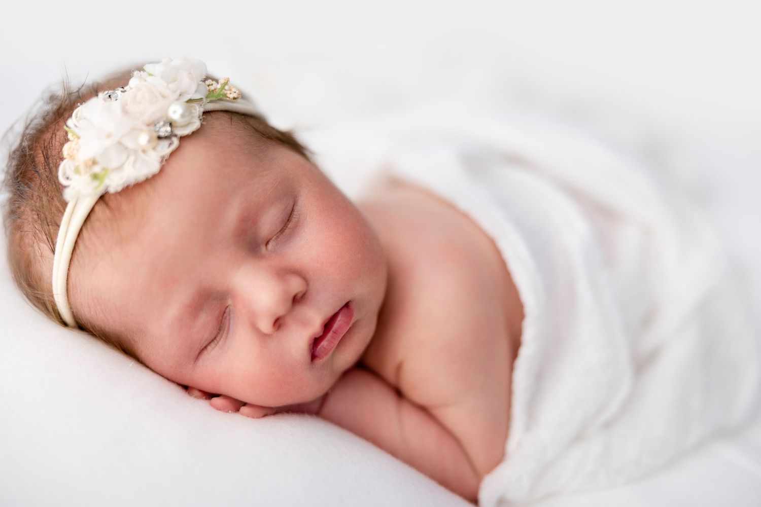 A sleeping baby wearing a white headband with a flower lies peacefully on white bedding.