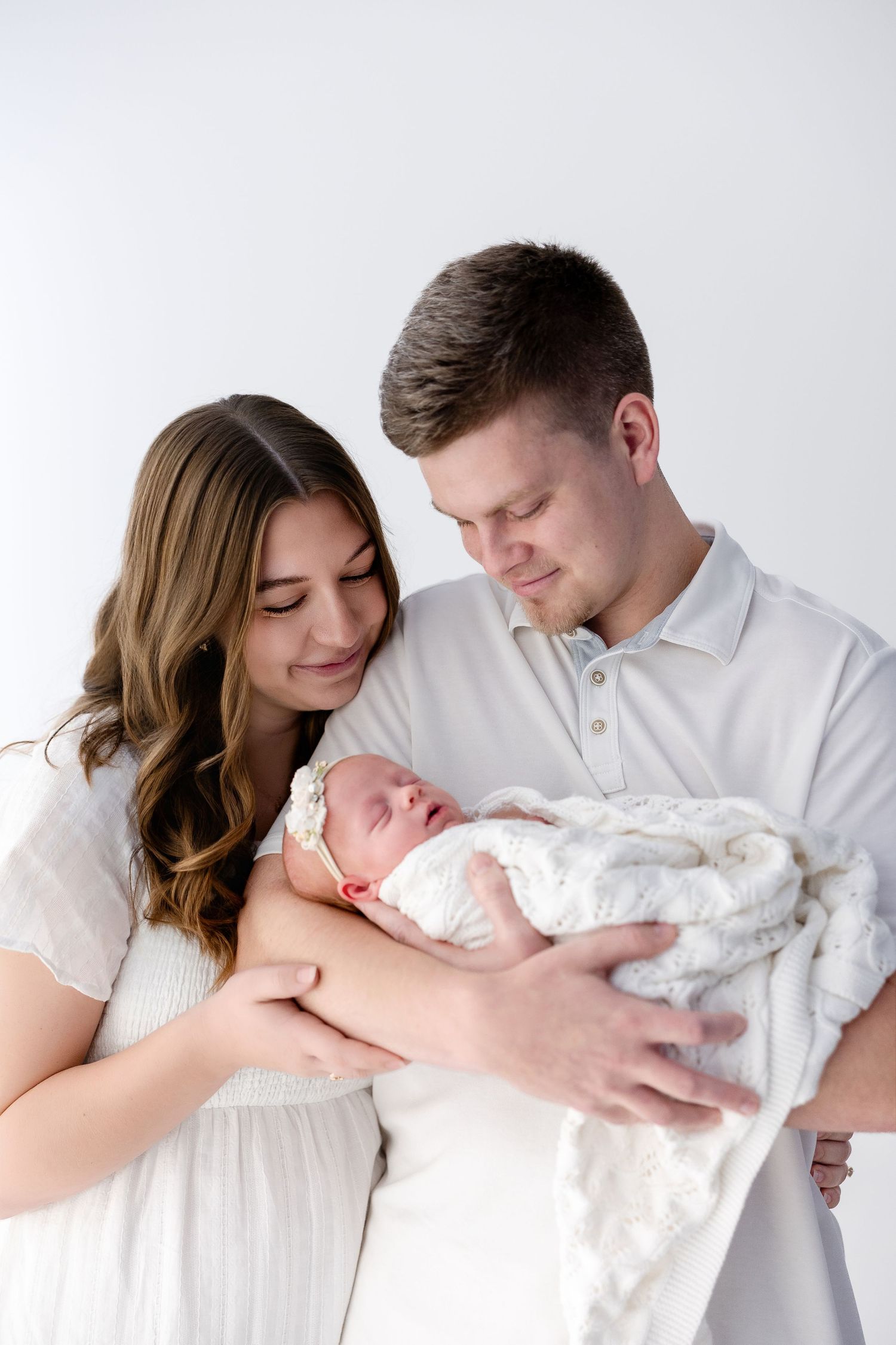 Parents in white clothing tenderly holding their newborn baby against minimalist backdrop.