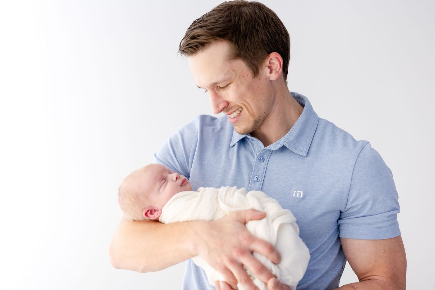 A person wearing a blue shirt holds a sleeping baby wrapped in white blankets.