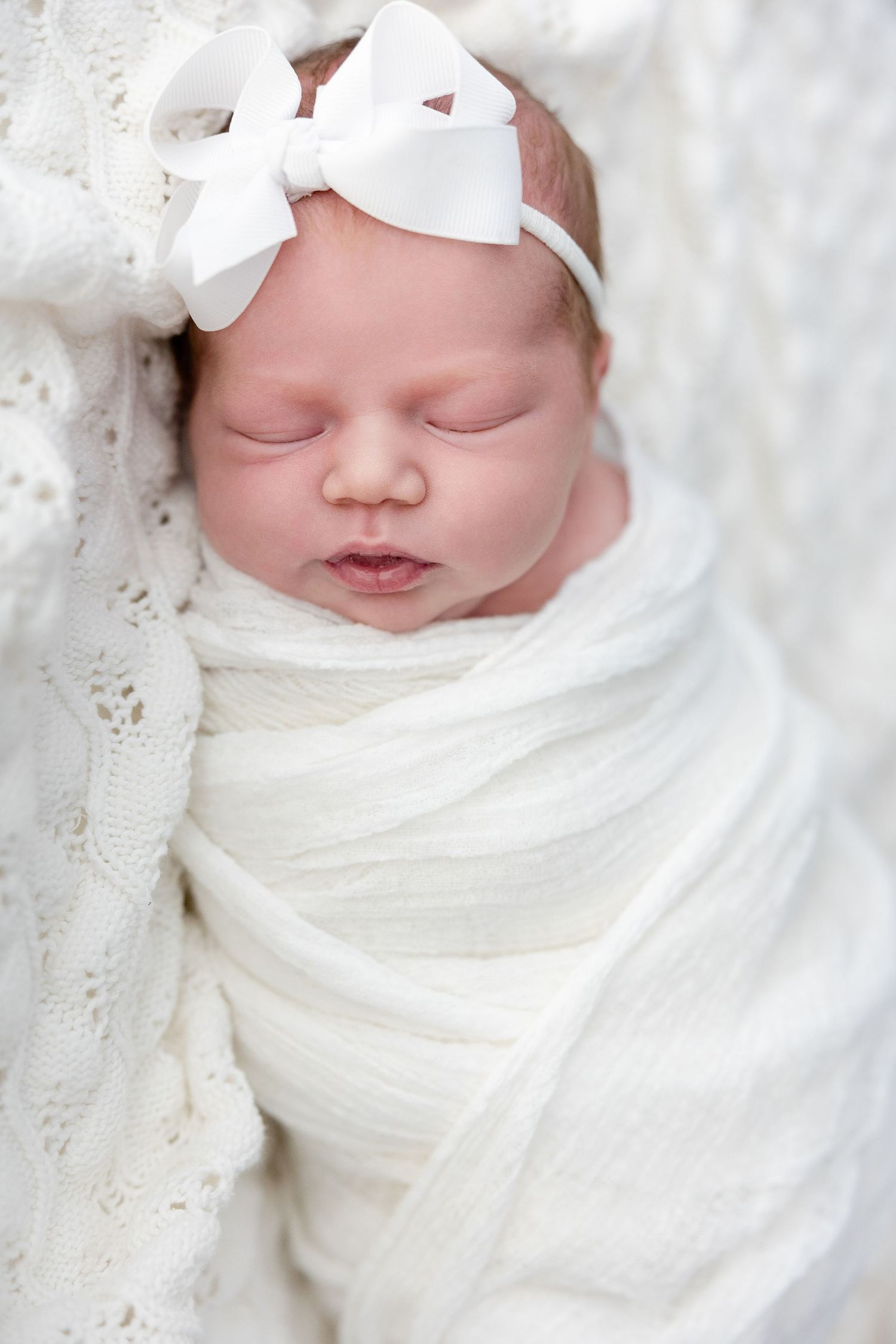 A sleeping newborn baby swaddled in white fabric wearing a white bow headband on soft textured blanket.