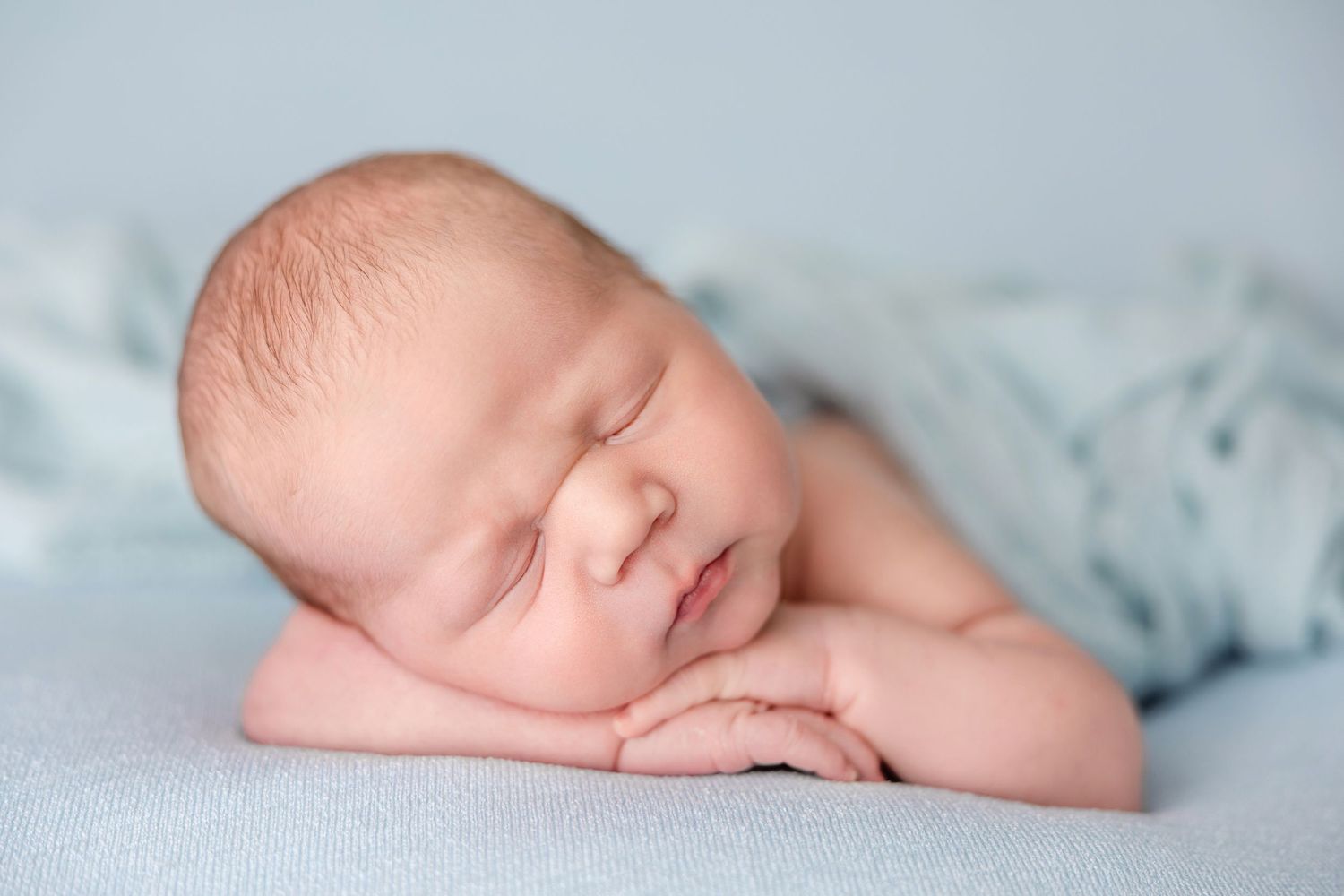 A sleeping newborn baby rests peacefully on soft blue fabric with gentle lighting in a studio portrait setting.