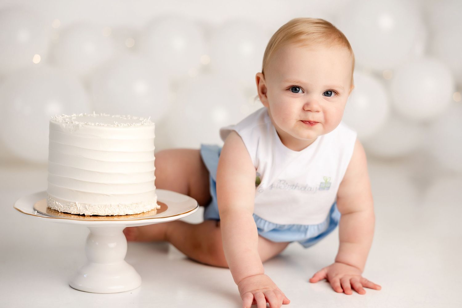 Baby in white outfit reaching for birthday cake on white background.