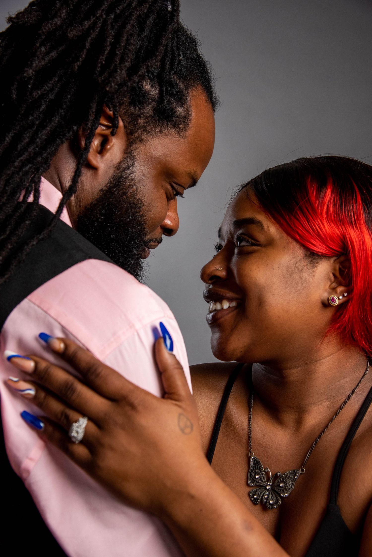 Intimate portrait of a couple embracing, with vibrant red hair and blue nails adding pops of color against a grey background.