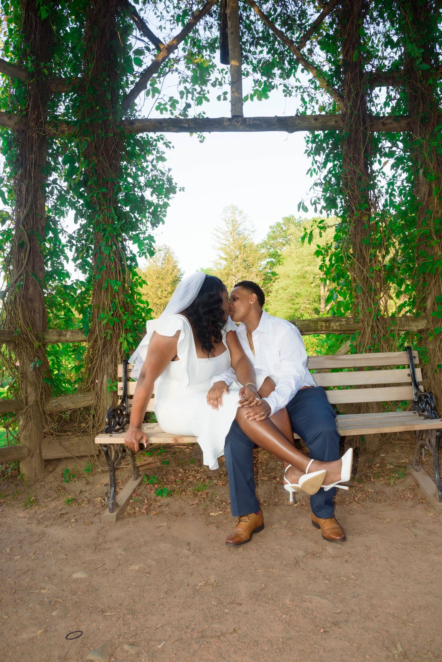 A romantic moment shared on a wooden bench surrounded by lush greenery and vines in a garden setting.