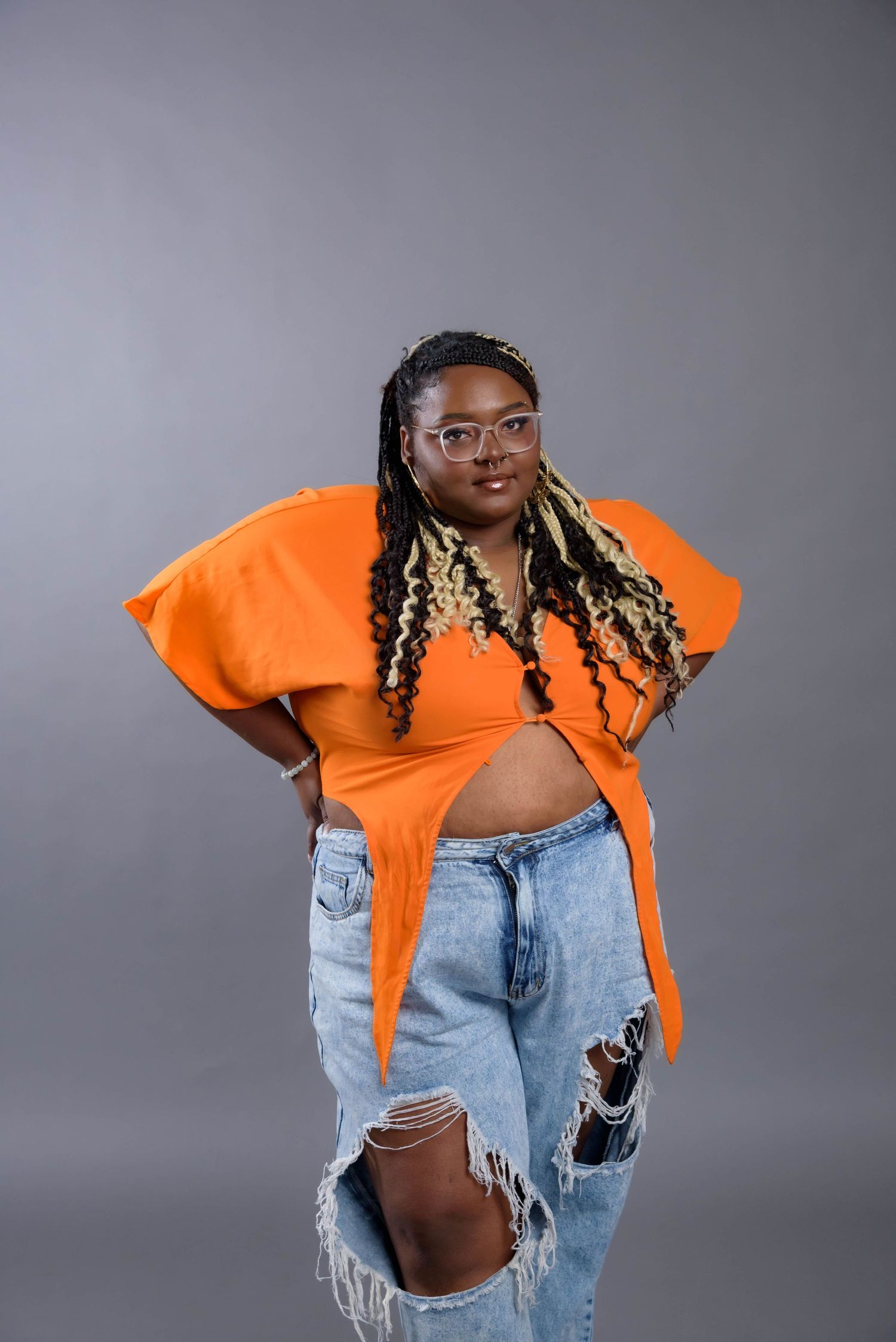 Fashion portrait of person in orange top and distressed jeans against grey background.
