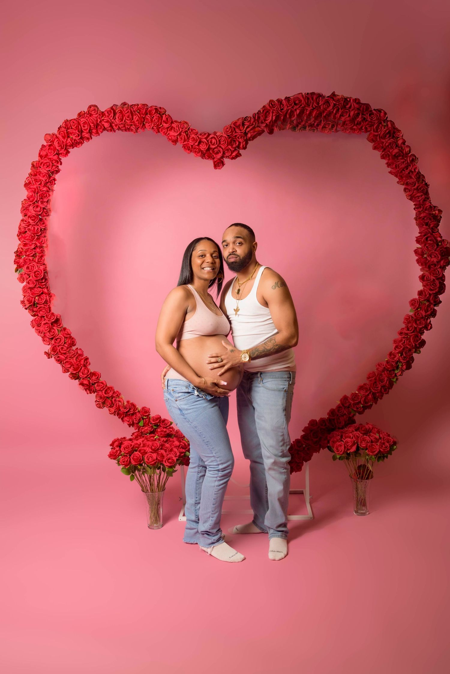 A maternity photo shoot with a red rose heart arch backdrop against a pink wall.