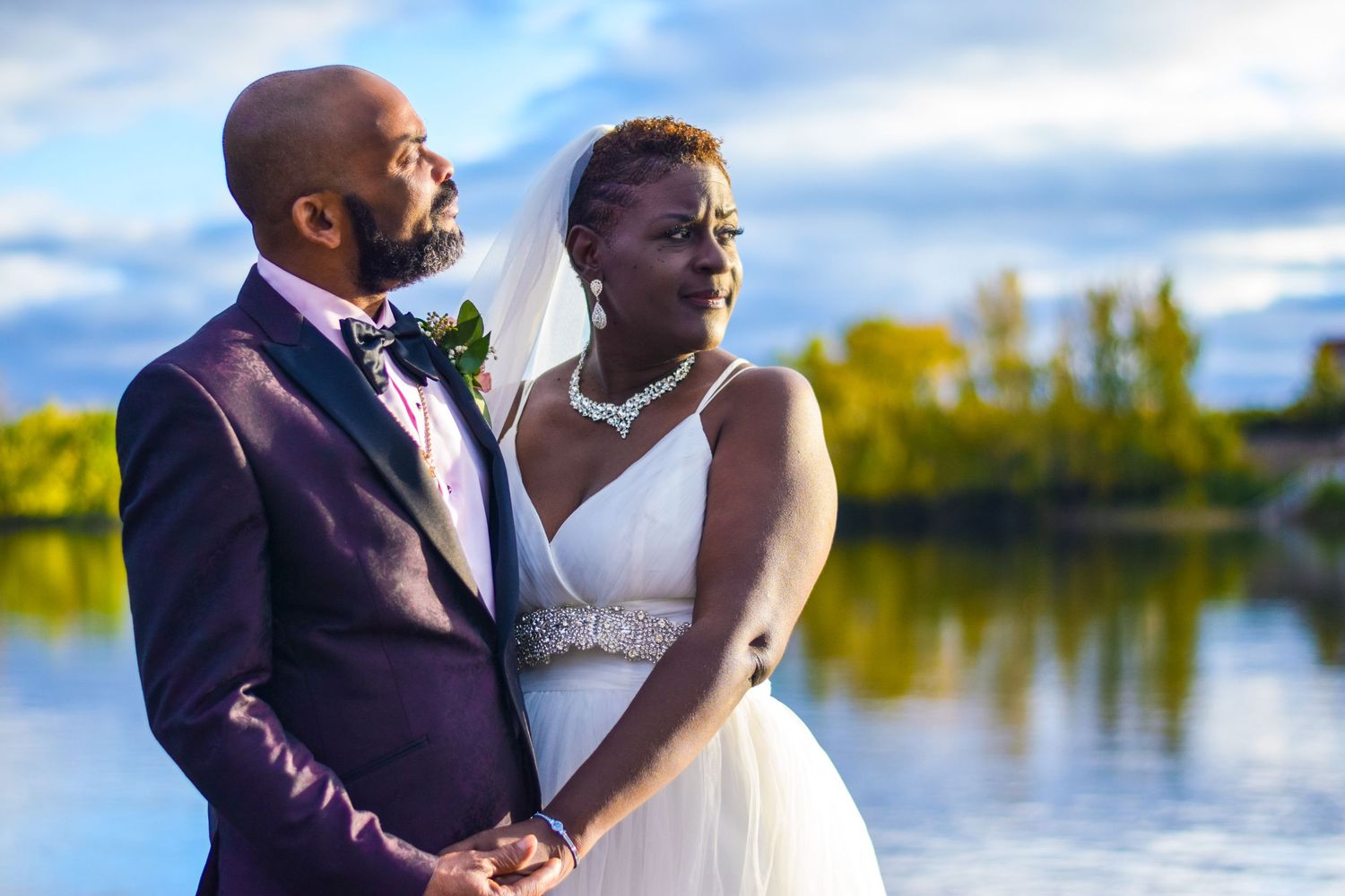 A couple in wedding attire stands by a serene lake during golden hour.