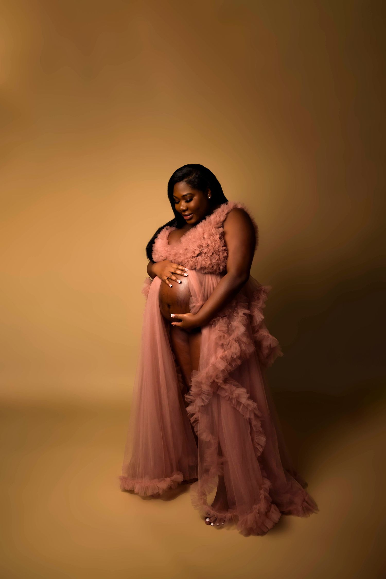 A maternity portrait in a flowing pink gown against a warm brown background.