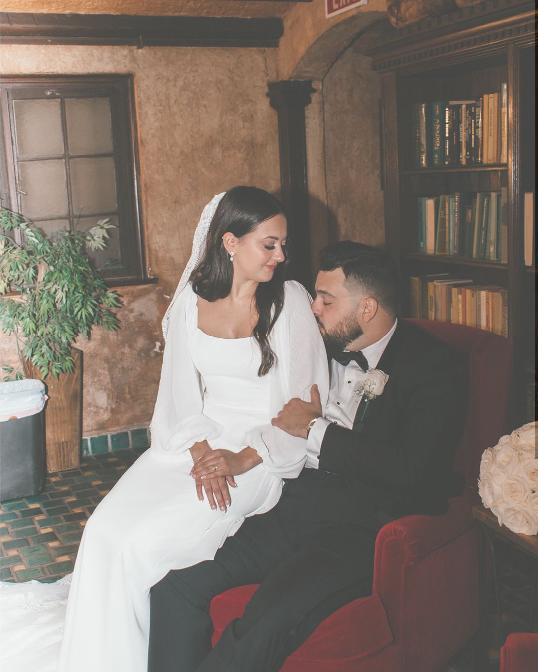 Newlyweds share tender moments on vintage red couch in rustic indoor setting.