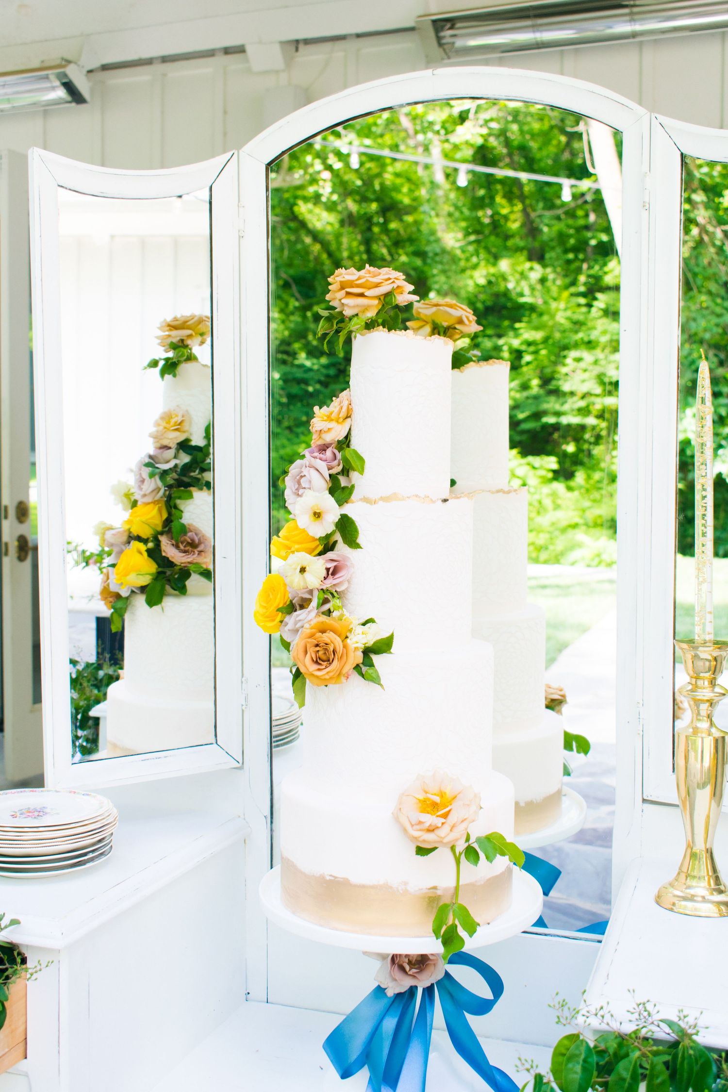 Elegant white tiered wedding cake with yellow and pink roses displayed in front of a mirror.