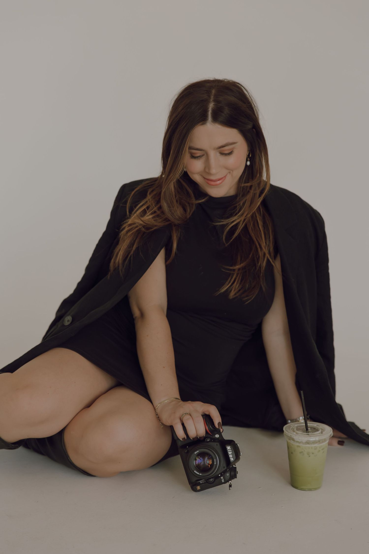 Photographer in black outfit sitting with camera and green drink on studio floor.