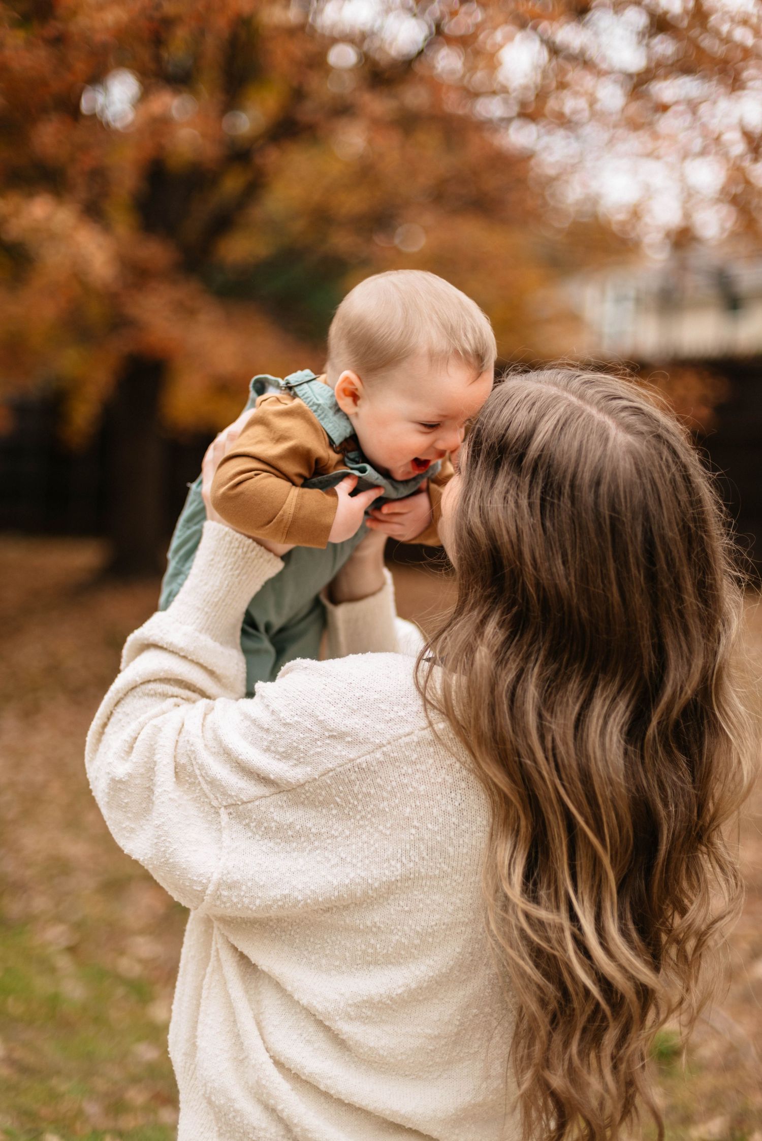 Mother in cream sweater playing with baby during autumn outdoor photoshoot.
