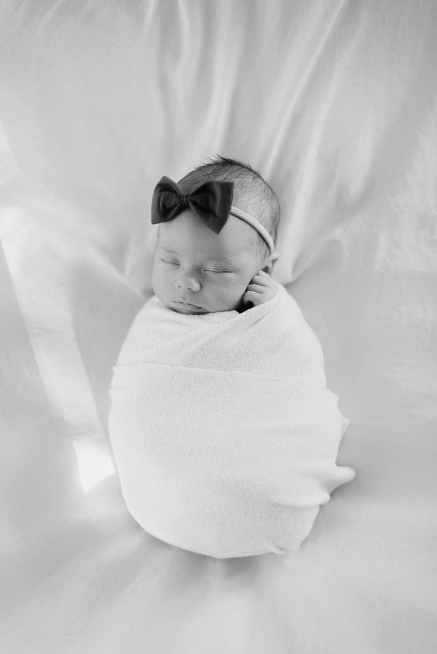 Peaceful sleeping newborn wrapped in white swaddle with bow accessory.