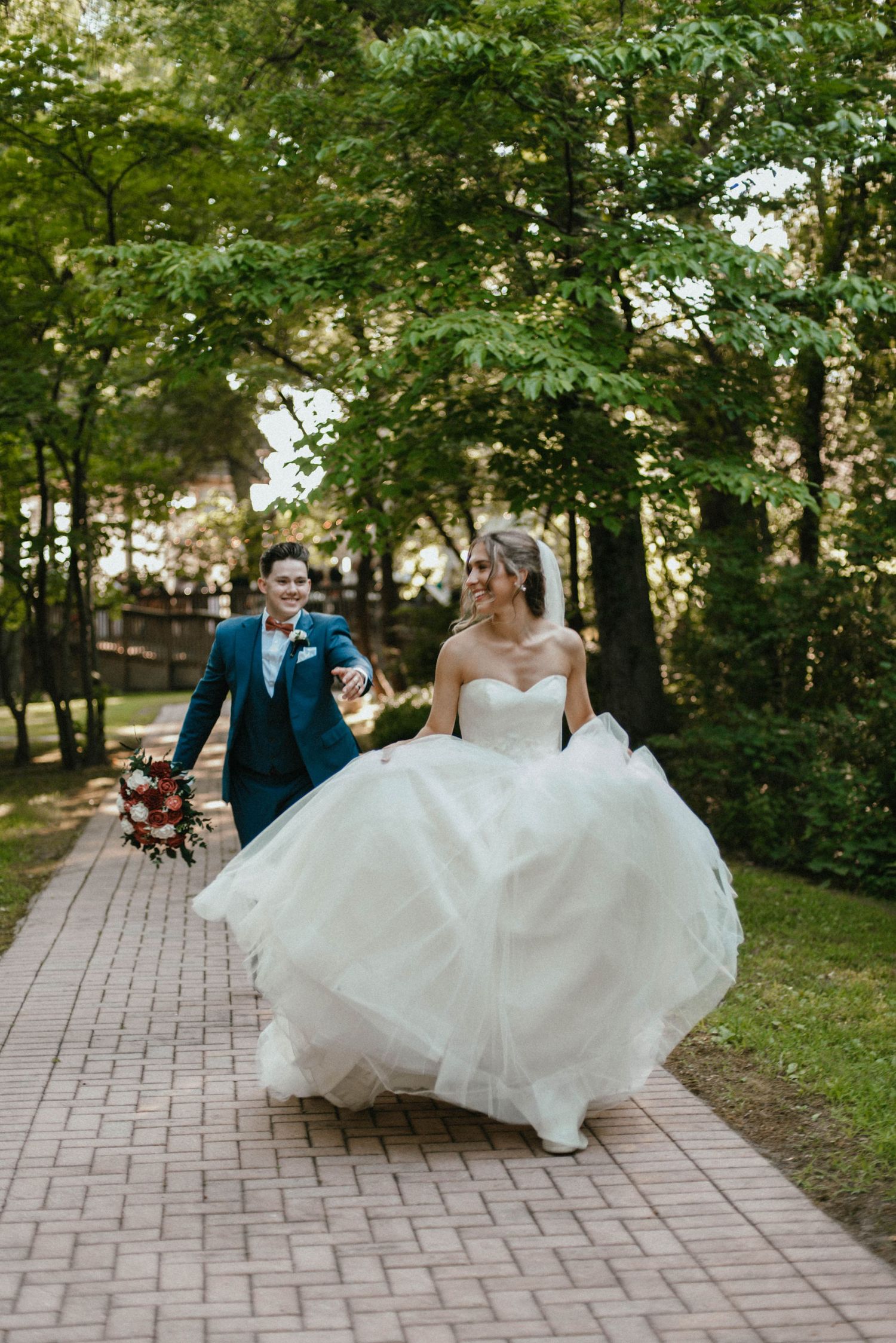White wedding dress flowing in motion along a brick pathway surrounded by green trees.