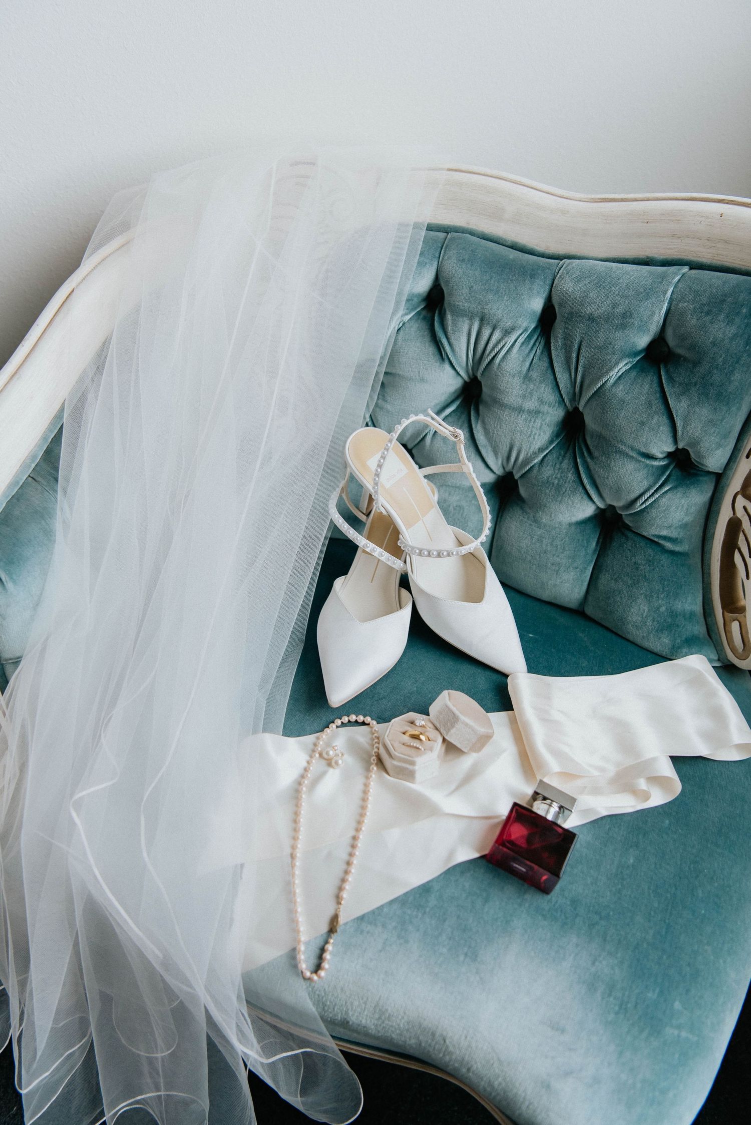 Wedding accessories including white shoes, veil, and pearl necklace arranged on a teal velvet chair.