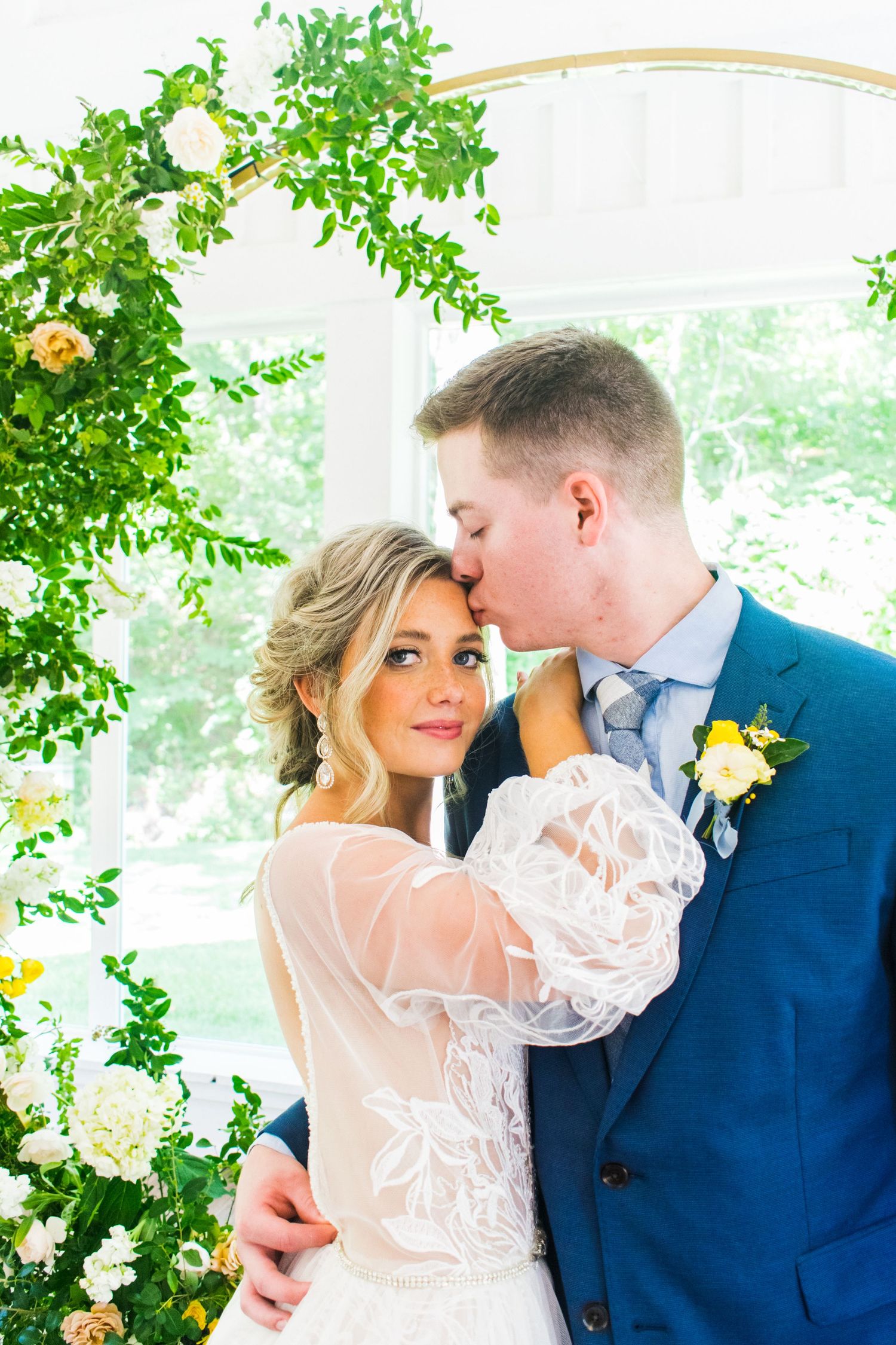 Wedding couple in blue suit and white lace dress embrace in front of floral greenery.