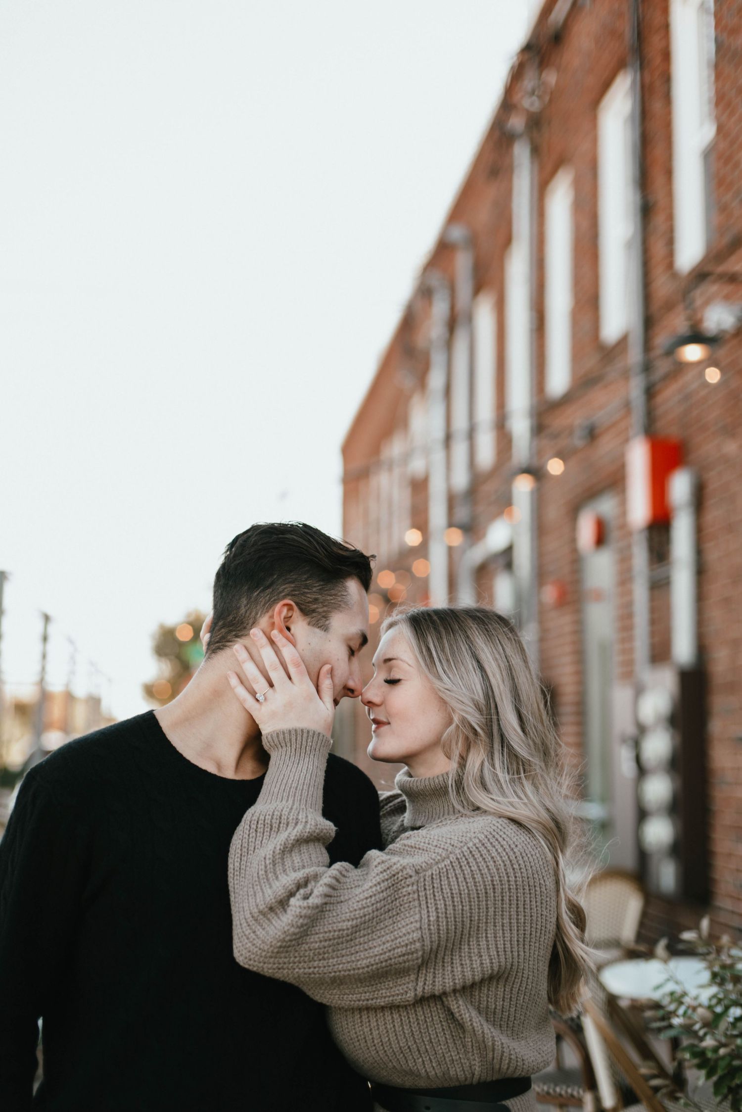 Romantic autumn engagement photoshoot against brick building with string lights.