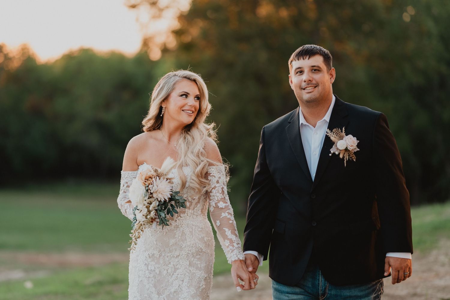 A bride and groom hold hands outdoors at golden hour, she in lace gown with bouquet, he in dark blazer with boutonniere.