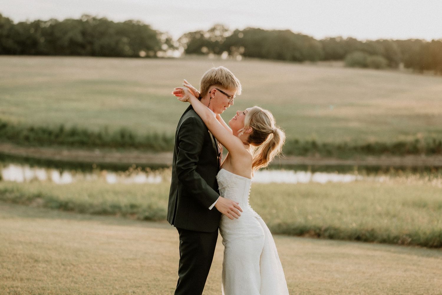 Bride and groom sharing a tender embrace in a golden sunlit field near a serene pond.