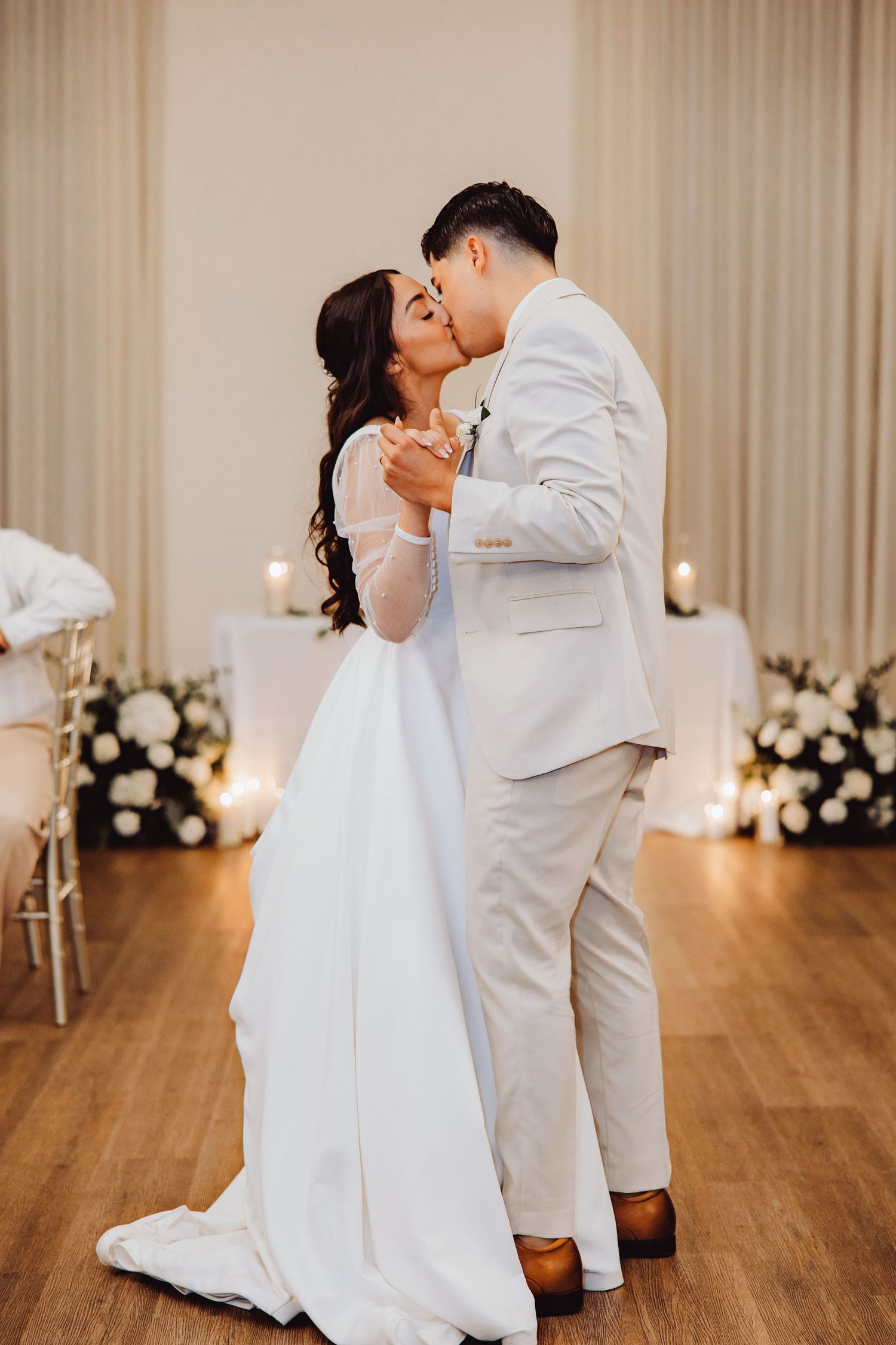 Newlyweds share their first dance kiss in an elegant white reception hall with floral arrangements and candlelight.
